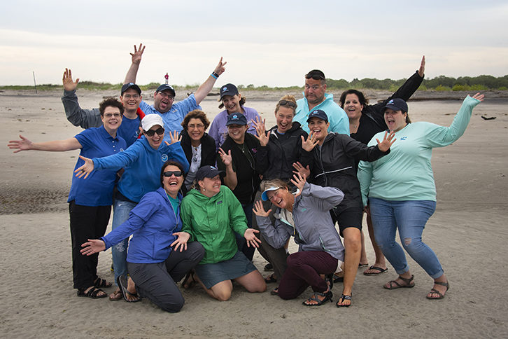 A group of 15 adults look excited and pose for a photo on a beach.