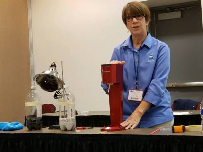 A woman in a blue shirt stands in front of a room and presents a science demonstration.
