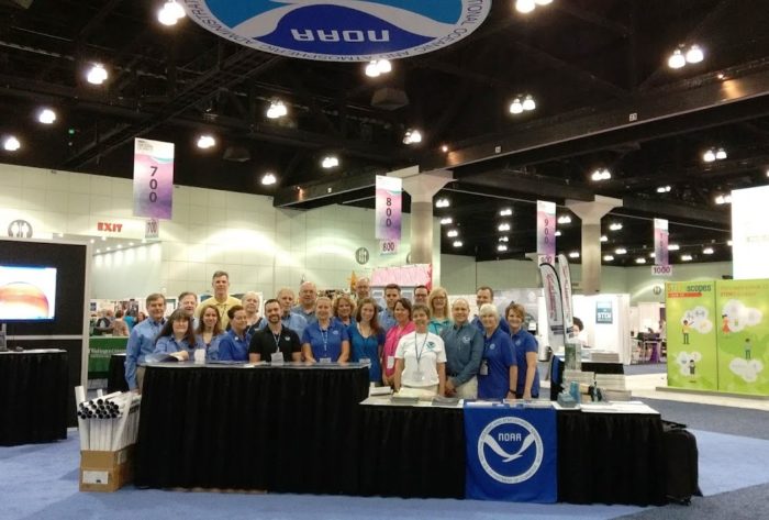 A group of people working at a booth in an exhibit hall and posing for a photo underneath a NOAA banner.