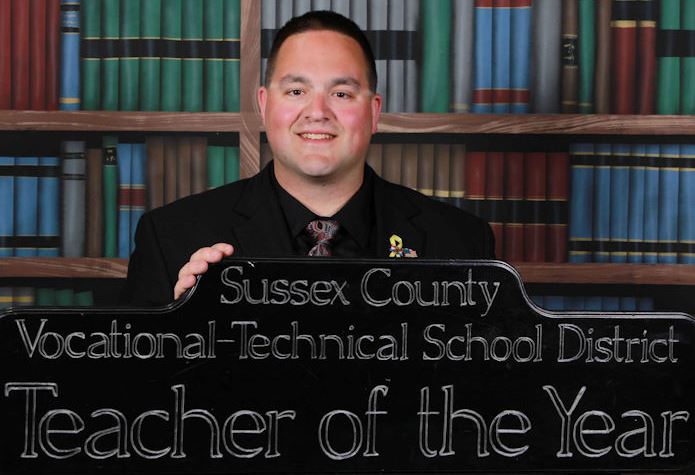 A man stands in front of a bookcase holding his Sussex County Vocational-Technical School District Teacher of the Year award.