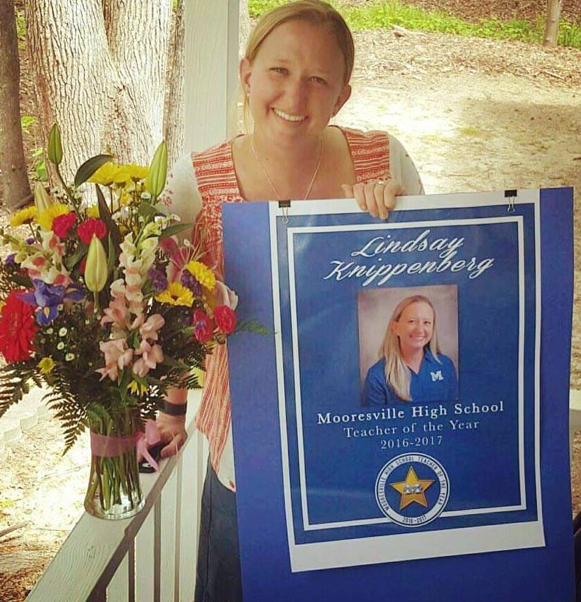 A woman standing outside next to a vase of flowers, holds a sign indicating she won the Mooresville High School Teacher of the Year 2016-2017.