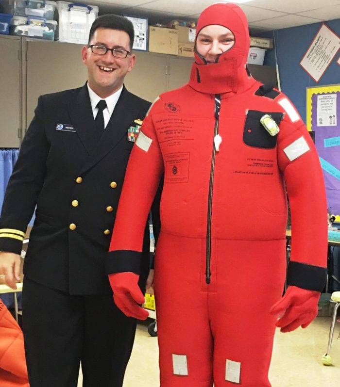 Two people standing next to each other. One in a military uniform and the other in a red, full body, water survival suit where you can only see their eyes and nose.