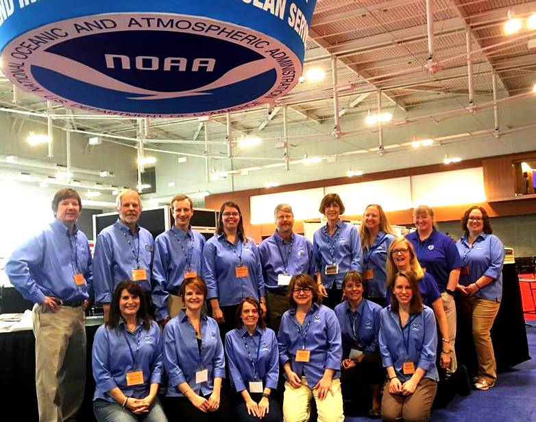 A group of men and women pose in front of the NOAA booth at the NSTA Conference. A large NOAA emblem hangs above them.