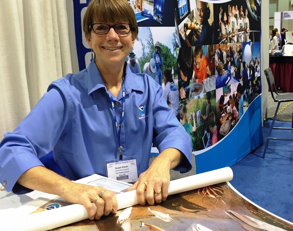 A woman sitting in front of a NOAA Education Booth banner, while she rolls up posters.