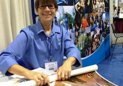 A woman sitting in front of a NOAA Education Booth banner, while she rolls up posters.