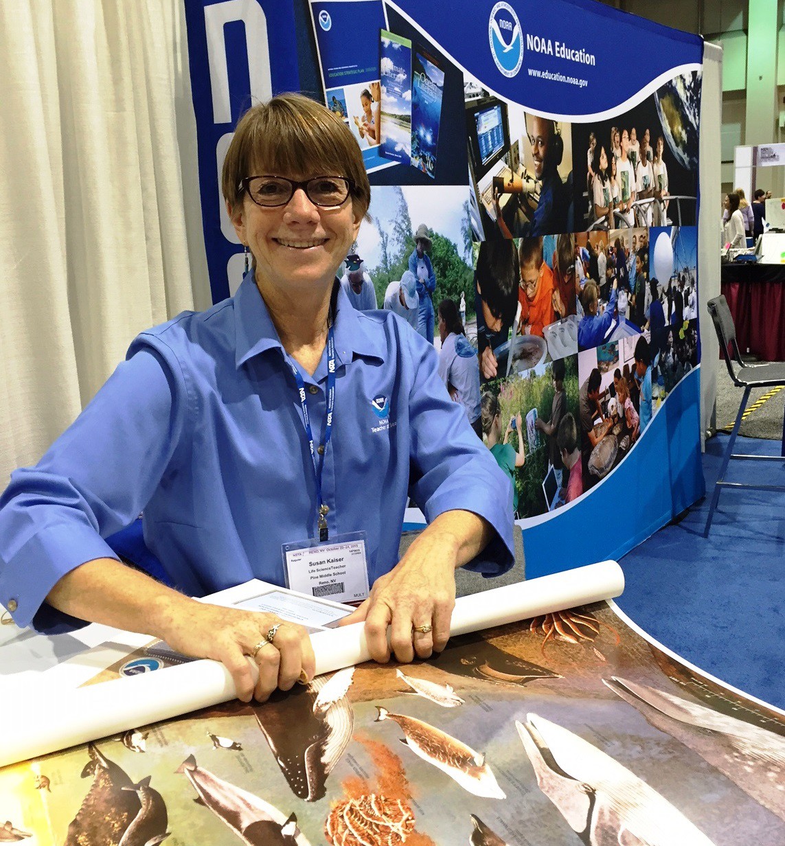 A woman sitting in front of a NOAA Education Booth banner, while she rolls up posters.