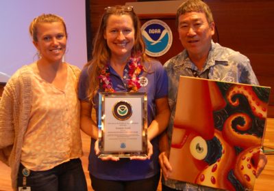 Two women and a man stand in front of NOAA signage. The center woman is holding an award while the man on the right is holding an art piece.