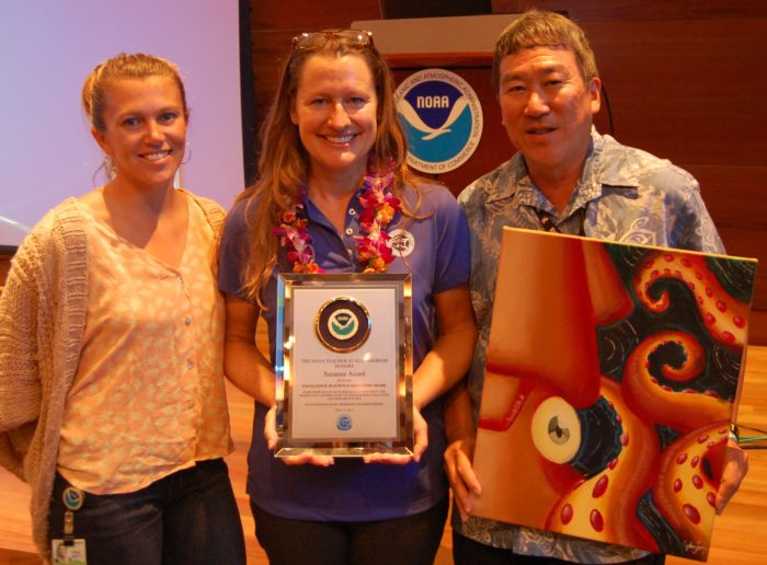 Two women and a man stand in front of NOAA signage. The center woman is holding an award while the man on the right is holding an art piece.
