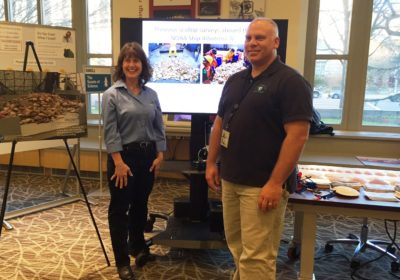 A woman is standing to the left by an easel with photos attached to it and a man is to her right. He is standing near a table with items on it and a screen with photos on it are in the background.