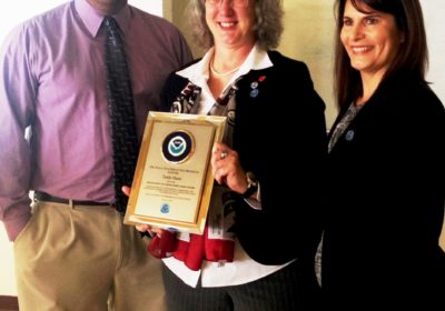 A man and two woman stand together while the woman in the center holds an award.