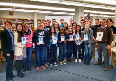 A group of students and two adults stand together in a library. The teacher is holding an award while some of the students are holding up signs.