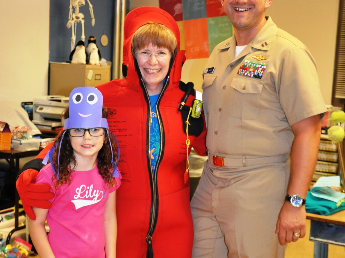 There are three people standing in a classroom. One is a child, the other a woman in a red, full body, survival suit and the last is a man in a military uniform.