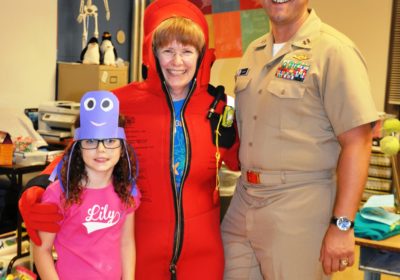 There are three people standing in a classroom. One is a child, the other a woman in a red, full body, survival suit and the last is a man in a military uniform.