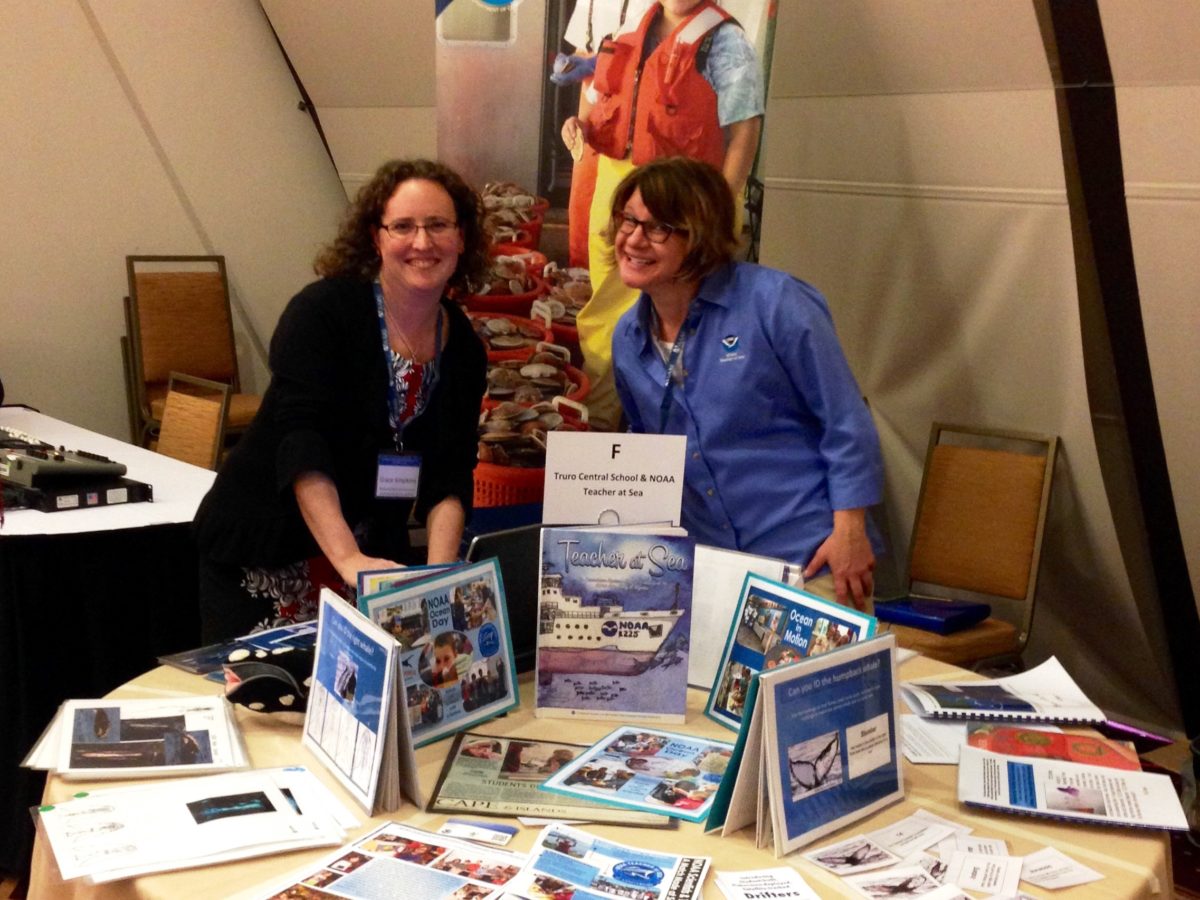 Two women stand in the middle of a NOAA Teacher at Sea sign behind them and a display table full of items in front of them.