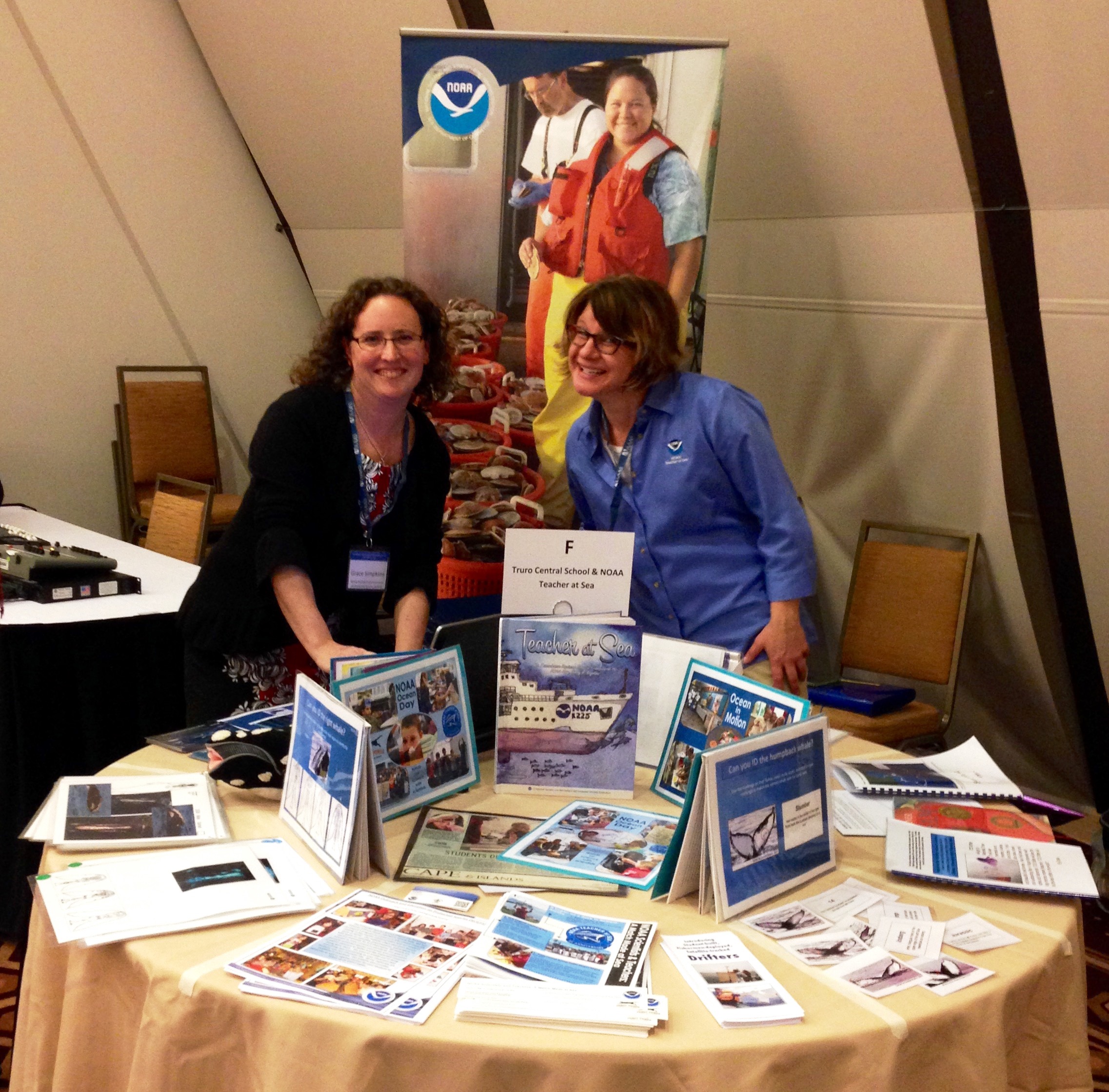Two women stand in the middle of a NOAA Teacher at Sea sign behind them and a display table full of items in front of them.