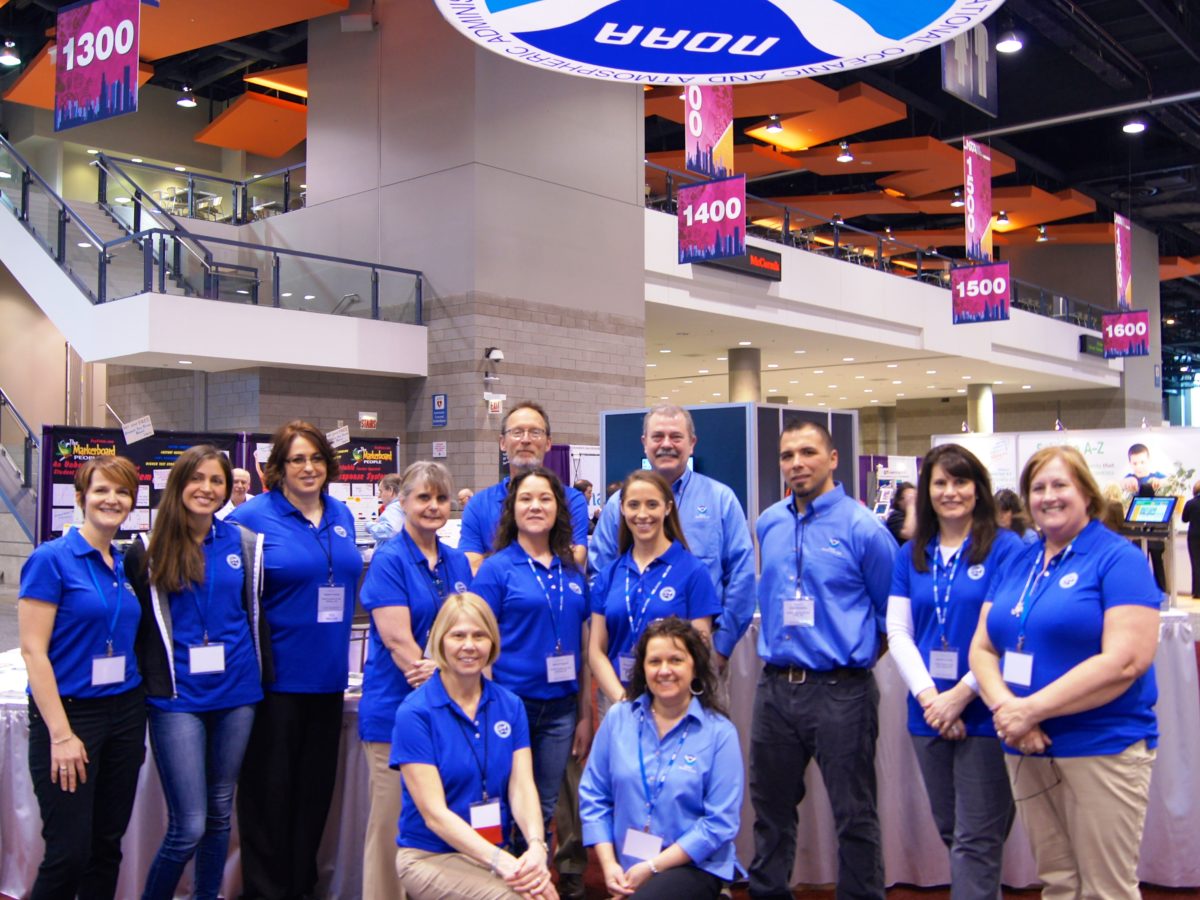 A group of men and woman stand in the NOAA booth in a large conference center. The NOAA logo is hanging above them.