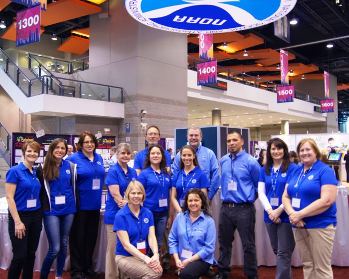 A group of men and woman stand in the NOAA booth in a large conference center. The NOAA logo is hanging above them.
