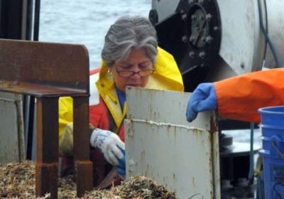 A woman dressed in rain gear and gloves is aboard a ship looking at something in her hand.