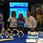 Two women and a man stand behind a table with shark items on it. There is NOAA signage in the background.