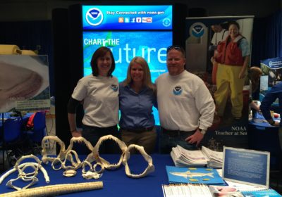 Two women and a man stand behind a table with shark items on it. There is NOAA signage in the background.