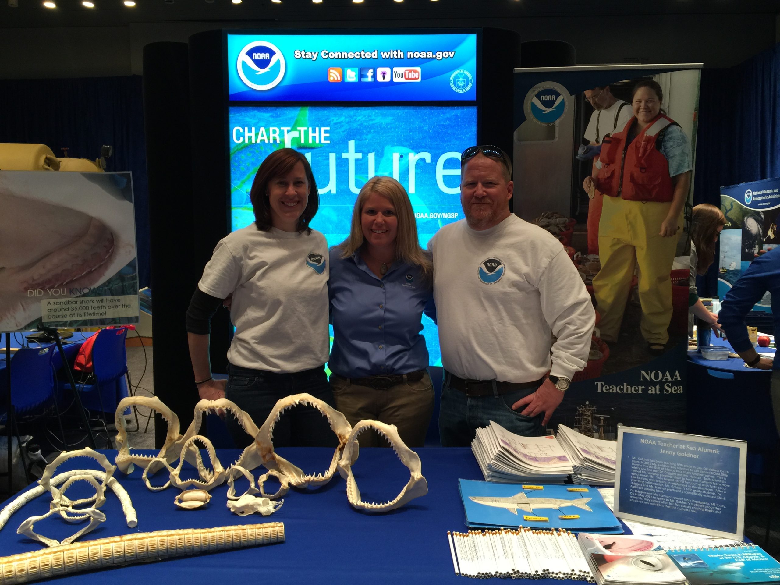 Two women and a man stand behind a table with shark items on it. There is NOAA signage in the background.