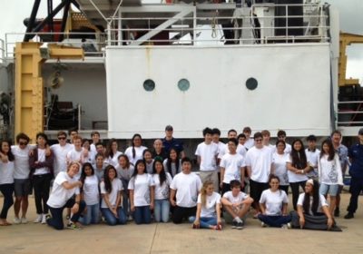 A group of students and adults gather on a pier with a ship in the background.