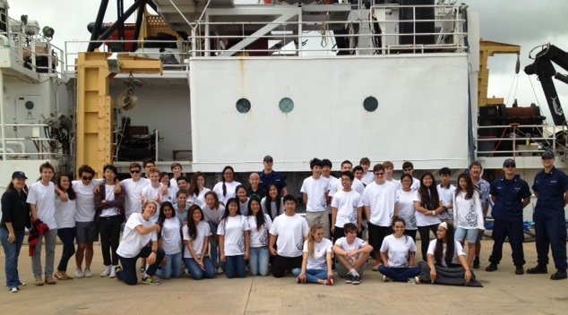 A group of students and adults gather on a pier with a ship in the background.
