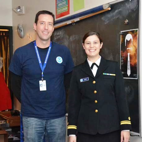 A man and a woman in a military uniform stand in a classroom.