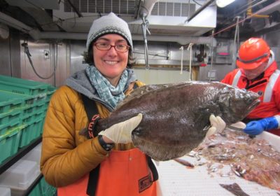 A woman stands holding a fish with a man in the background looking at other fish on a conveyor.