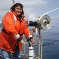 A woman standing on board a ship with water in the background.