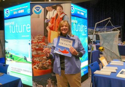 A woman stands in front of a NOAA sign holding a Fishing for Facts sign.