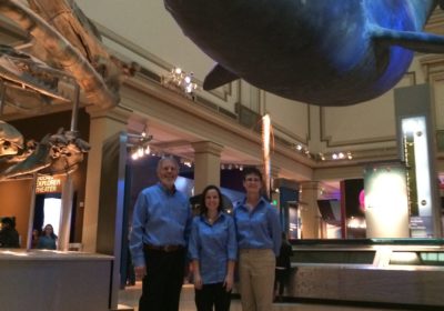 A man and two women stand in a large room with a large sea animal above them.
