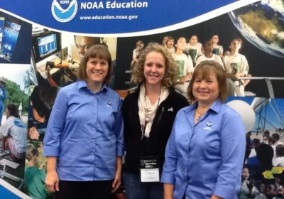 Three women stand in front of a NOAA Education sign.