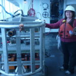 A woman stands next to a TowCam aboard a ship with the ocean off to the right.