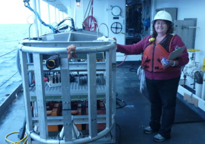 A woman stands next to a TowCam aboard a ship with the ocean off to the right.