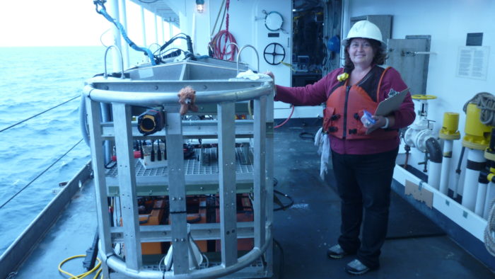 A woman stands next to a TowCam aboard a ship with the ocean off to the right.