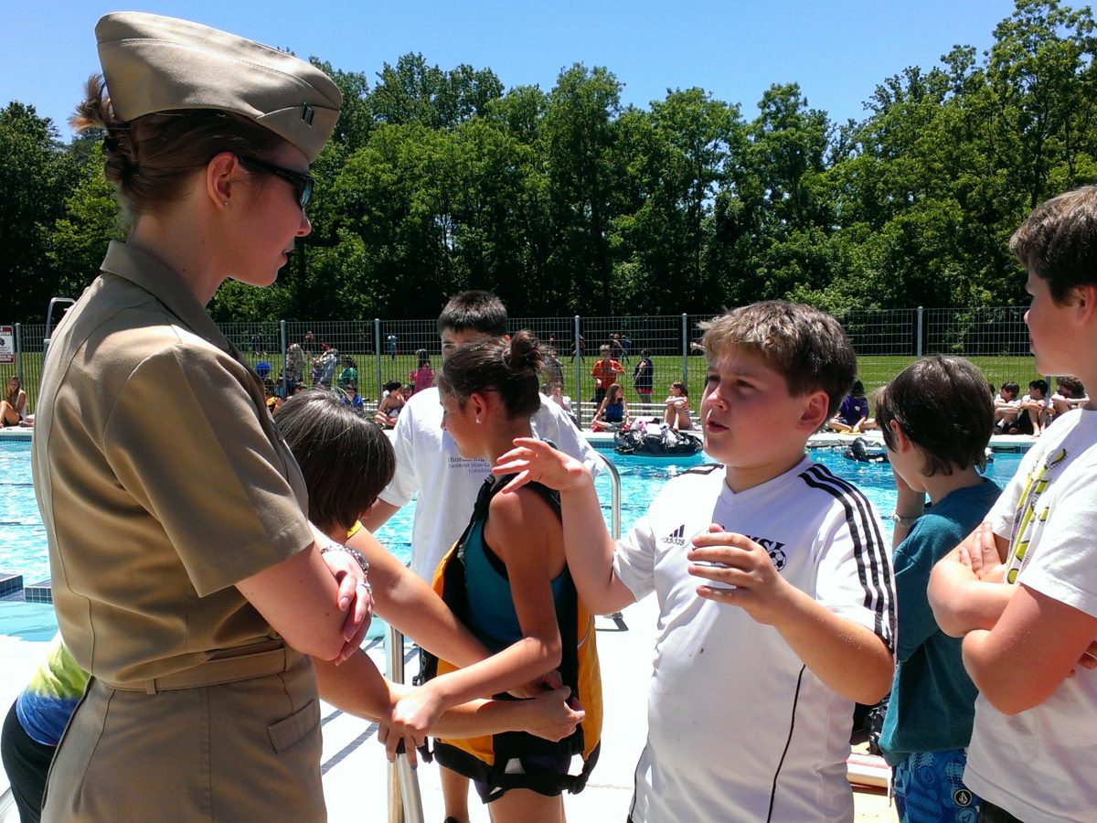 A woman in a military uniform is speaking to a group of students in front of an outdoor pool.
