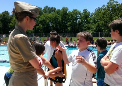 A woman in a military uniform is speaking to a group of students in front of an outdoor pool.