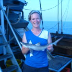 A woman on a ship holding a shark with water in the background.