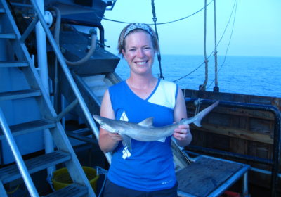 A woman on a ship holding a shark with water in the background.