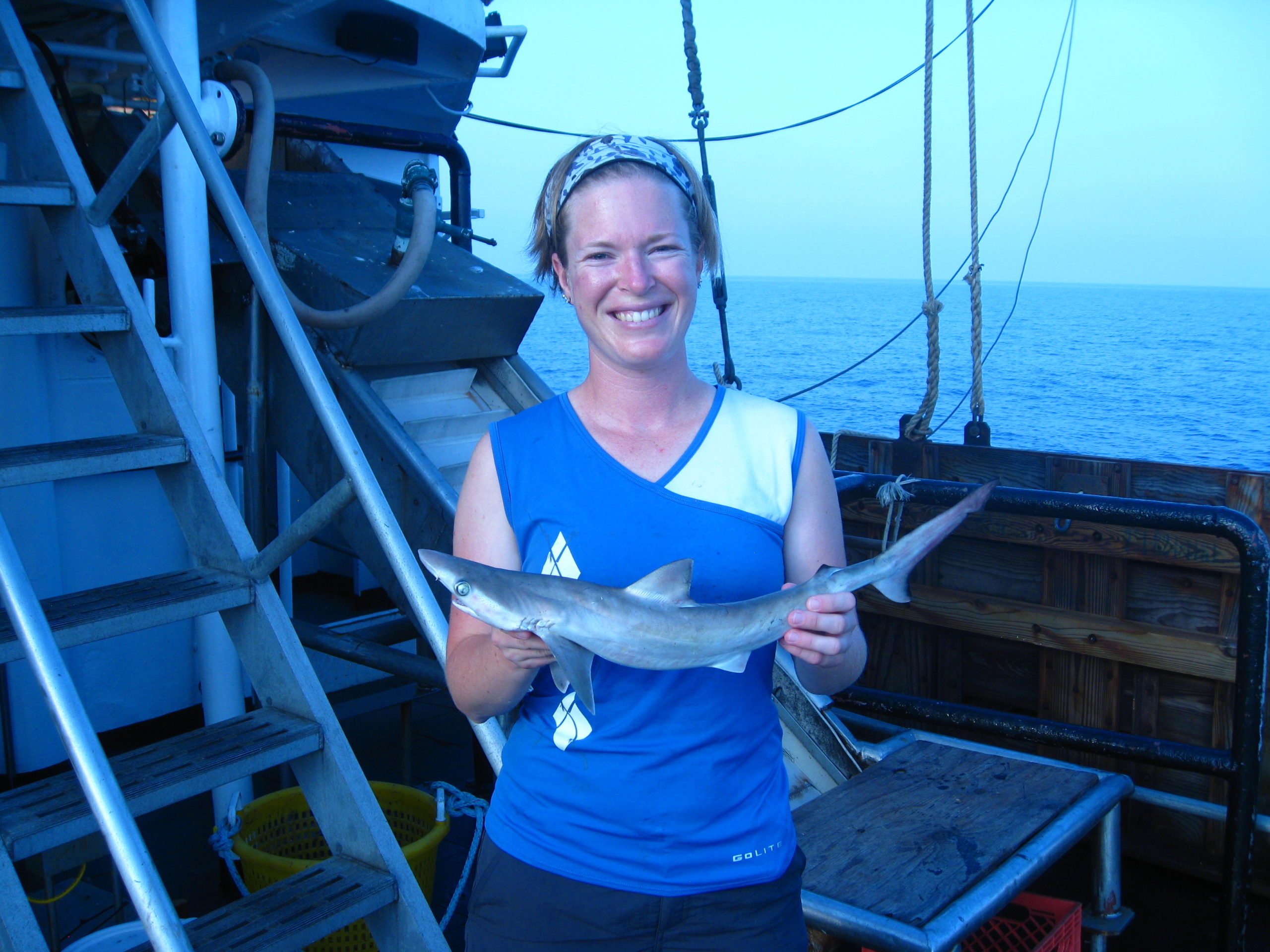 A woman on a ship holding a shark with water in the background.