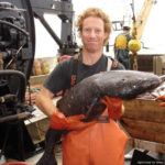 A man on a ship holding a fish with fish and another man in the background.