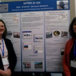 Two women stand in front of a sign that says "Sisters at Sea".