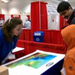 A woman talks with people at a booth in a conference hall.