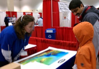 A woman talks with people at a booth in a conference hall.