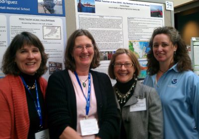 Four women stand in front of a NOAA Teacher at Sea sign.
