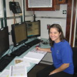 A woman sits at a desk with three computer monitors and an open notebook.