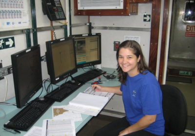 A woman sits at a desk with three computer monitors and an open notebook.