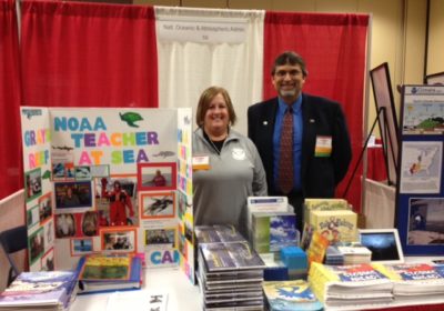 A woman and a man stand behind a NOAA Teacher at Sea display table.