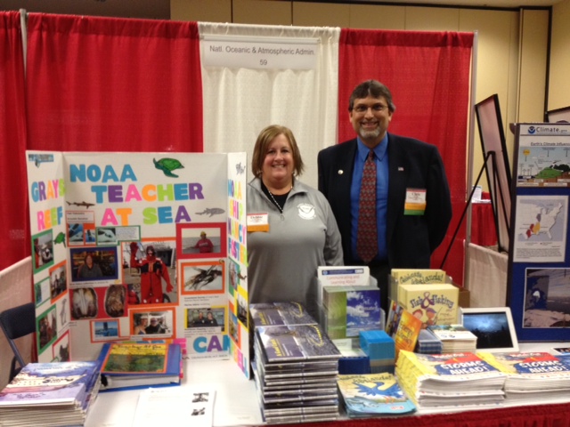A woman and a man stand behind a NOAA Teacher at Sea display table.